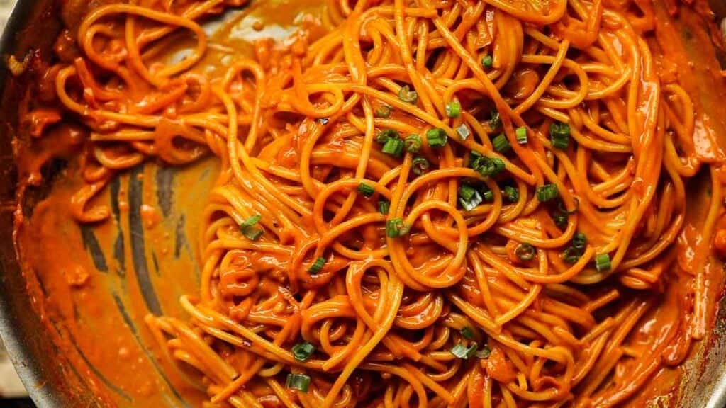 A close-up of vegan gochujang vodka pasta tossed in a rich tomato sauce with a garnish of chopped green herbs in a pan. The sauce is thick and clings to the pasta, with some visible streaks on the sides of the pan.